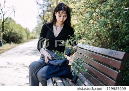 Young woman sits on park bench while packing tomato seedlings into bag. Potted vegetable plants, city dweller gardening, organic food growing. Young woman sits on park bench while packing tomato seedlings into bag. Potted vegetable plants, city dweller gardening, organic food growing. 139983009