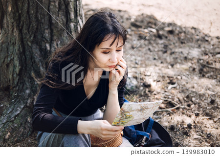 Person sits under tree while carefully reading paper map in forest. Reading paper map, finding the way, lost in the woods concept, Route planning strategy. Person sits under tree while carefully reading paper map in forest. Reading paper map, finding the way, lost in the woods concept, Route planning strategy. 139983010