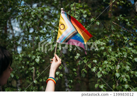 Hand holds inclusive pride flag against foliage. lgbtq diversity and inclusion, corporate pride support, lgbtq friendly business. Hand holds inclusive pride flag against foliage. lgbtq diversity and inclusion, corporate pride support, lgbtq friendly business. 139983012