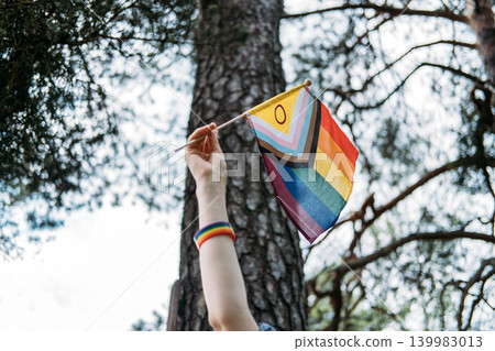 Hand raises progress pride flag next to tree trunk in forest. Human rights activism, support for LGBTQ community, non-binary person lifestyle. Hand raises progress pride flag next to tree trunk in forest. Human rights activism, support for LGBTQ community, non-binary person lifestyle. 139983013
