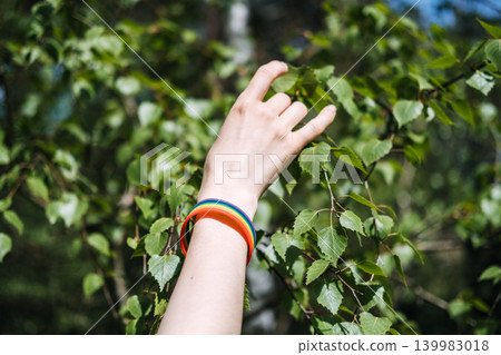 Hand wearing rainbow bracelet reaches toward green birch leaves in forest. Human rights organization, volunteering for LGBTQ cause, community support program. Hand wearing rainbow bracelet reaches toward green birch leaves in forest. Human rights organization, volunteering for LGBTQ cause, community support program. 139983018