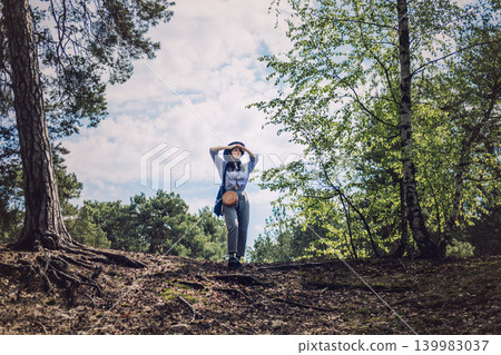 Woman stands on forest hill shielding eyes with hands while looking toward horizon. Eco friendly vacation, sustainable travel choices, responsible tourism, Leaving no trace. Woman stands on forest hill shielding eyes with hands while looking toward horizon. Eco friendly vacation, sustainable travel choices, responsible tourism, Leaving no trace. 139983037