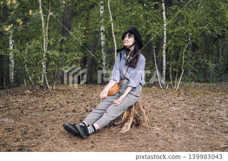 Person wearing hat sits on tree stump in forest with eyes closed and hands resting on lap. Forest bathing therapy, mental health outdoors, stress relief in nature. Person wearing hat sits on tree stump in forest with eyes closed and hands resting on lap. Forest bathing therapy, mental health outdoors, stress relief in nature. 139983043