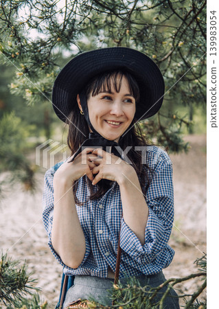 Woman wearing hat smiles under pine tree branches in forest. Solace in nature, forest bathing therapy, mindfulness in the woods. Woman wearing hat smiles under pine tree branches in forest. Solace in nature, forest bathing therapy, mindfulness in the woods. 139983054