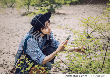 Woman crouches while examining plant with smartphone in sandy woodland area. Using technology for education, real-time nature sharing, mobile botanical guide. Woman crouches while examining plant with smartphone in sandy woodland area. Using technology for education, real-time nature sharing, mobile botanical guide. 139983060