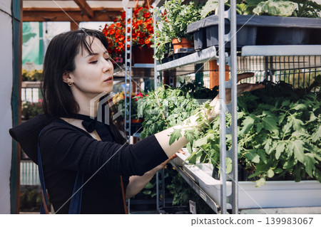 Woman selects leafy green vegetable plants from metal shelf at outdoor garden market. Food self-sufficiency, homegrown produce movement, urban agriculture lifestyle. Woman selects leafy green vegetable plants from metal shelf at outdoor garden market. Food self-sufficiency, homegrown produce movement, urban agriculture lifestyle. 139983067