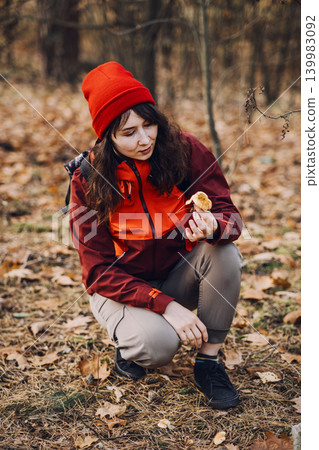 Woman crouches in forest and examines wild mushroom. wild mushroom foraging, gathering edible plants, sustainable self-sufficiency. Woman crouches in forest and examines wild mushroom. wild mushroom foraging, gathering edible plants, sustainable self-sufficiency. 139983092