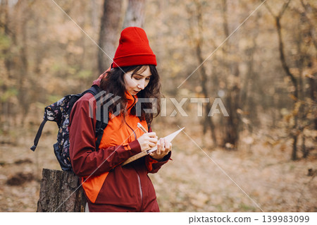 Woman stands in autumn forest and writes in notebook while leaning on tree stump. Working from anywhere, freelance outdoor workspace, nomadic professional life. Woman stands in autumn forest and writes in notebook while leaning on tree stump. Working from anywhere, freelance outdoor workspace, nomadic professional life. 139983099