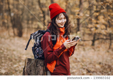Smiling woman looks at smartphone screen while leaning on tree stump in autumn forest. AI-driven wilderness assistance, mobile tools for trail safety, digital outdoor survival guide. Smiling woman looks at smartphone screen while leaning on tree stump in autumn forest. AI-driven wilderness assistance, mobile tools for trail safety, digital outdoor survival guide. 139983102