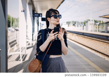 Young woman holds smartphone while standing on sunny railway platform. Modern railway commute, public transport network, sustainable urban travel, Waiting for train. Young woman holds smartphone while standing on sunny railway platform. Modern railway commute, public transport network, sustainable urban travel, Waiting for train. 139983109