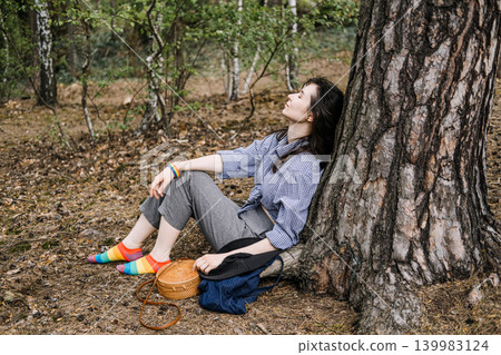 Person sits on ground leaning against tree in forest with eyes closed. mental wellness awareness, emotional support lgbt, forest bathing therapy. 139983124
