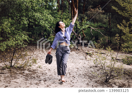 Barefoot woman stands on sand in forest with arm raised holding shoes. Nature therapy for stress relief, mental health awareness, emotional wellness at nature. 139983125