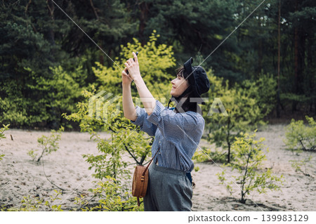 Woman stands in sandy forest clearing while taking photo with smartphone. Digital nomad leisure, balancing technology and nature, peaceful forest walk. 139983129
