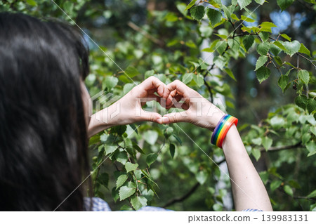 Woman makes heart gesture with hands wearing rainbow bracelet during pride month. Pride month, LGBTQ community, inclusive love, diversity representation. 139983131