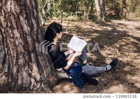 Young woman sits against tree trunk while reading book in forest. Mental health awareness, mindfulness practice, emotional wellbeing, Stress relief outdoor. 139983136