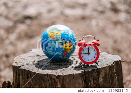 Small globe and red alarm clock sit on weathered tree stump in outdoor setting. Climate change urgency, global warming deadline, earth hour concept. Small globe and red alarm clock sit on weathered tree stump in outdoor setting. Climate change urgency, global warming deadline, earth hour concept. 139983145