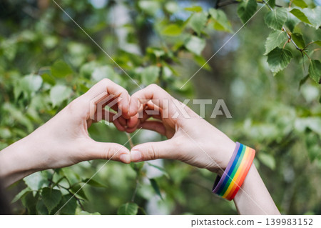 Hands form heart shape against green foliage with rainbow bracelet on wrist. Pride month celebration, LGBTQ pride support, queer community solidarity. Hands form heart shape against green foliage with rainbow bracelet on wrist. Pride month celebration, LGBTQ pride support, queer community solidarity. 139983152