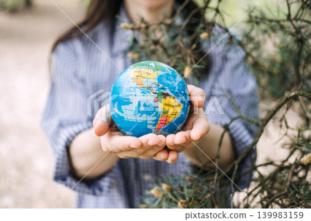 Person holds small globe with both hands outdoors near pine branches. Human touch for nature, protecting global ecosystem, Earth day celebration. 139983159