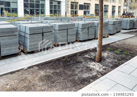 Large stacks of granite paving slabs sit on wooden pallets at urban street construction site. Granite paving slabs, stone paver installation, bulk construction materials. Large stacks of granite paving slabs sit on wooden pallets at urban street construction site. Granite paving slabs, stone paver installation, bulk construction materials. 139983293