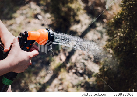 Hands hold hose nozzle while spraying water on small evergreen bushes. Eco-friendly home maintenance, water saving technology, outdoor water management. Hands hold hose nozzle while spraying water on small evergreen bushes. Eco-friendly home maintenance, water saving technology, outdoor water management. 139983305