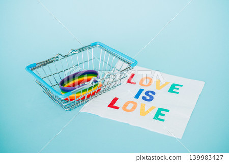 Rainbow silicone bracelet sits inside miniature metal shopping basket next to LOVE IS LOVE napkin. LGBTQ Subscription box diversity, ethical gifting guide, rainbow merchandise kit. 139983427