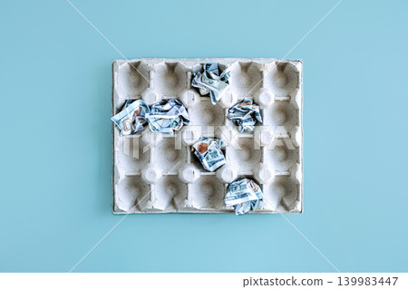 Crumpled US dollar banknotes rest in empty cardboard egg tray on blue surface. Household expenses growth, grocery shopping budget, expensive food staples. Crumpled US dollar banknotes rest in empty cardboard egg tray on blue surface. Household expenses growth, grocery shopping budget, expensive food staples. 139983447