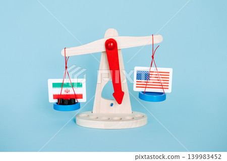 Wooden scales weighing Iranian flag with barrels against United States flag on blue background. Middle Eastern conflict impact, regional power balance, Strait of Hormuz security. Wooden scales weighing Iranian flag with barrels against United States flag on blue background. Middle Eastern conflict impact, regional power balance, Strait of Hormuz security. 139983452