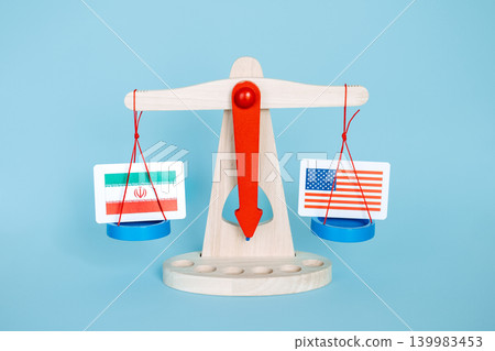 Wooden scales weighing Iranian flag against United States flag on blue background. Middle Eastern conflict impact, regional power balance, Strait of Hormuz security. Wooden scales weighing Iranian flag against United States flag on blue background. Middle Eastern conflict impact, regional power balance, Strait of Hormuz security. 139983453