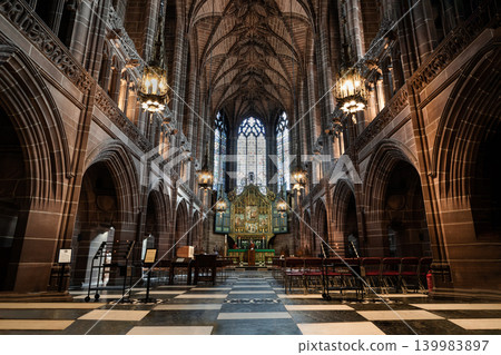 Warm light from hanging lanterns illuminates the towering stone arches and intricate vaulted ceiling of a grand, historic cathedral, casting a serene glow down a checkered aisle toward a magnificent Warm light from hanging lanterns illuminates the towering stone arches and intricate vaulted ceiling of a grand, historic cathedral, casting a serene glow down a checkered aisle toward a magnificent 139983897