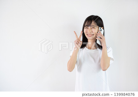 A young Japanese woman makes a peace sign while talking on her smartphone in front of a white background. A young Japanese woman makes a peace sign while talking on her smartphone in front of a white background. 139984076