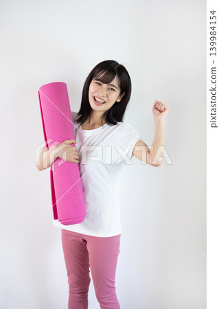 A young Japanese woman smiles, holding a yoga mat, in front of a white background. A young Japanese woman smiles, holding a yoga mat, in front of a white background. 139984154