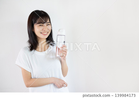 A young Japanese woman smiling with a water bottle in her hand in front of a white background. A young Japanese woman smiling with a water bottle in her hand in front of a white background. 139984173