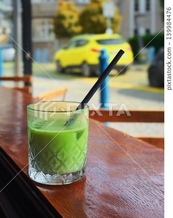 Iced matcha latte in patterned glass with straw stands on wooden cafe table by window with city street blur, copy space. Concept of matcha lifestyle, slow living, cafe break, wellness drink Iced matcha latte in patterned glass with straw stands on wooden cafe table by window with city street blur, copy space. Concept of matcha lifestyle, slow living, cafe break, wellness drink 139984476