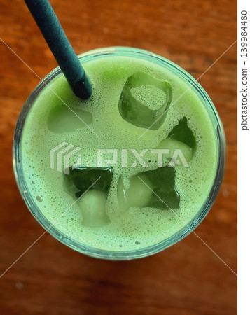 Top view of foamy matcha drink with ice cubes and craft straw in glass on wooden table, green bubbles and chilled surface. Concept of matcha texture, cafe refreshment, wellness beverage trend Top view of foamy matcha drink with ice cubes and craft straw in glass on wooden table, green bubbles and chilled surface. Concept of matcha texture, cafe refreshment, wellness beverage trend 139984480