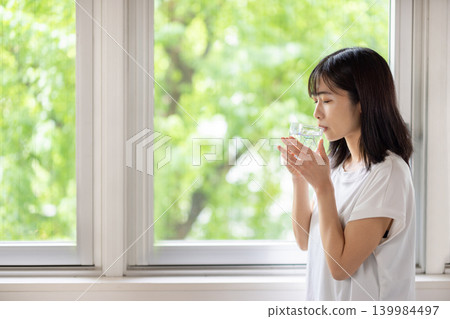 A young Japanese woman holds a glass of water and looks out the window. A young Japanese woman holds a glass of water and looks out the window. 139984497