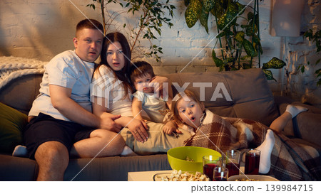Family watching movie at home with popcorn and red drinks on cozy sofa. Family watching movie at home with popcorn and red drinks on cozy sofa. 139984715