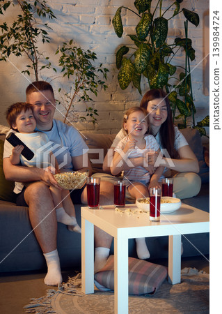 Cheerful family enjoying movie night with popcorn in cozy living room. Cheerful family enjoying movie night with popcorn in cozy living room. 139984724