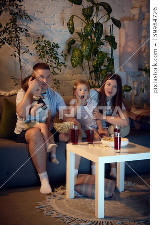 Family watching suspenseful movie on sofa with popcorn and berry drinks. Family watching suspenseful movie on sofa with popcorn and berry drinks. 139984726