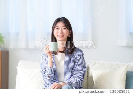 A smiling Japanese woman drinking coffee on a sofa. 139985931