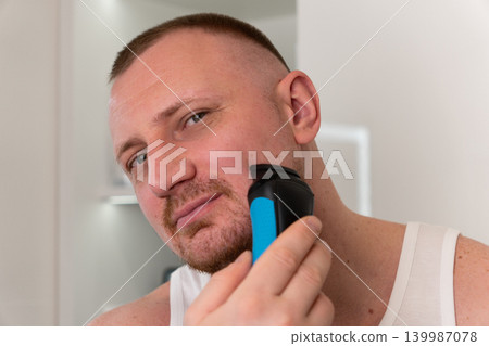 Handsome young man shaving his face with electric shaver in bathroom mirror during morning routine. Male grooming, skincare and personal care concept. Handsome young man shaving his face with electric shaver in bathroom mirror during morning routine. Male grooming, skincare and personal care concept. 139987078