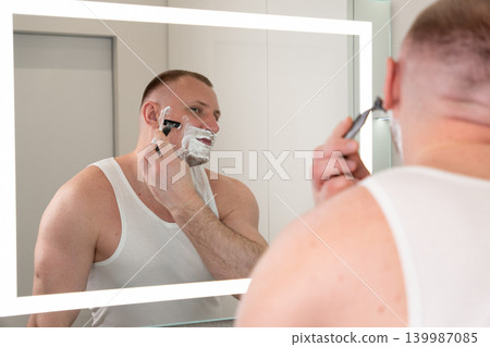 Handsome young man shaving his face with razor in bathroom mirror during morning routine. Male grooming, skincare and personal care concept. Handsome young man shaving his face with razor in bathroom mirror during morning routine. Male grooming, skincare and personal care concept. 139987085