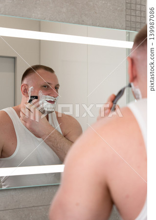 Vertical close up portrait of handsome young man shaving his face with razor in bathroom mirror during morning routine. Male grooming, skincare and personal care concept. Vertical close up portrait of handsome young man shaving his face with razor in bathroom mirror during morning routine. Male grooming, skincare and personal care concept. 139987086