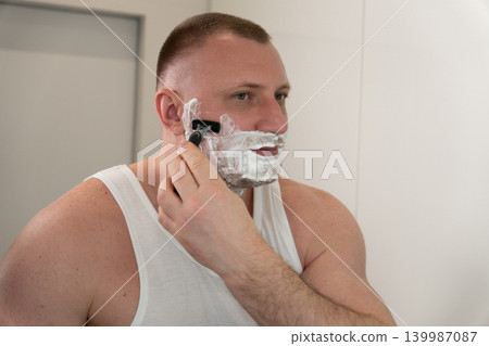 Handsome young man shaving his face with razor in bathroom mirror during morning routine. Male grooming, skincare and personal care concept. Handsome young man shaving his face with razor in bathroom mirror during morning routine. Male grooming, skincare and personal care concept. 139987087