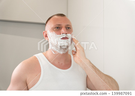 Close up portrait of handsome young man applying shaving foam on face in bathroom mirror. Morning male grooming and skincare routine concept before shaving. Close up portrait of handsome young man applying shaving foam on face in bathroom mirror. Morning male grooming and skincare routine concept before shaving. 139987091