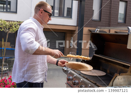 A man prepares pizza on a gas grill on a sunny outdoor terrace. The scene shows a modern lifestyle cooking moment with a focus on grilling food outdoors. Perfect for concepts of summer cooking A man prepares pizza on a gas grill on a sunny outdoor terrace. The scene shows a modern lifestyle cooking moment with a focus on grilling food outdoors. Perfect for concepts of summer cooking 139987121