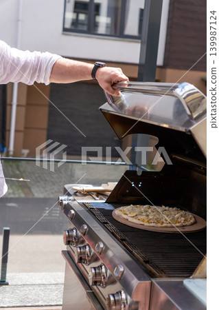 Vertical shot of a man preparing pizza on a gas grill on an outdoor terrace. The image captures a modern lifestyle cooking scene with focus on outdoor grilling, summer atmosphere, and homemade food Vertical shot of a man preparing pizza on a gas grill on an outdoor terrace. The image captures a modern lifestyle cooking scene with focus on outdoor grilling, summer atmosphere, and homemade food 139987124