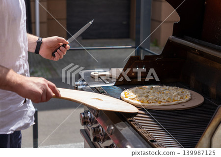 A man prepares pizza on a gas grill on a sunny outdoor terrace. The scene shows a modern lifestyle cooking moment with a focus on grilling food outdoors. Perfect for concepts of summer cooking 139987125