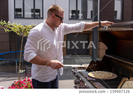 A man prepares pizza on a gas grill on a sunny outdoor terrace. The scene shows a modern lifestyle cooking moment with a focus on grilling food outdoors. Perfect for concepts of summer cooking A man prepares pizza on a gas grill on a sunny outdoor terrace. The scene shows a modern lifestyle cooking moment with a focus on grilling food outdoors. Perfect for concepts of summer cooking 139987126