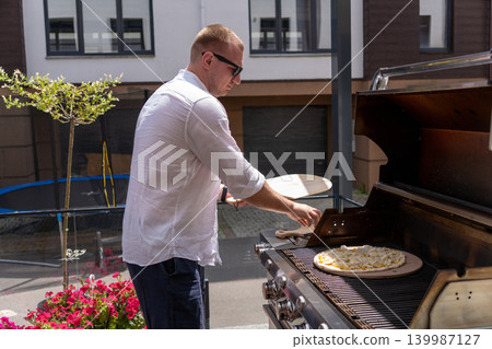 A man prepares pizza on a gas grill on a sunny outdoor terrace. The scene shows a modern lifestyle cooking moment with a focus on grilling food outdoors. Perfect for concepts of summer cooking A man prepares pizza on a gas grill on a sunny outdoor terrace. The scene shows a modern lifestyle cooking moment with a focus on grilling food outdoors. Perfect for concepts of summer cooking 139987127