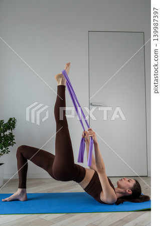 Vertical portrait of a fit young woman performing stretching and fitness exercises with resistance band at home. Female training indoors in sportswear during wellness and healthy lifestyle routine Vertical portrait of a fit young woman performing stretching and fitness exercises with resistance band at home. Female training indoors in sportswear during wellness and healthy lifestyle routine 139987397